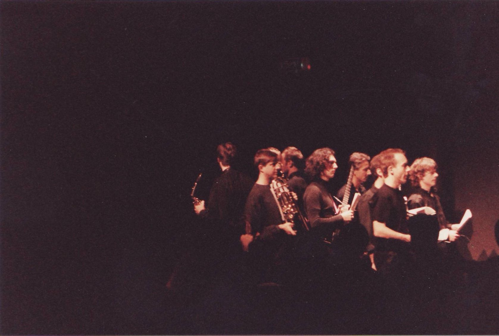 A group of people standing on a stage, lit with red lighting. They hold papers, likely performing.
