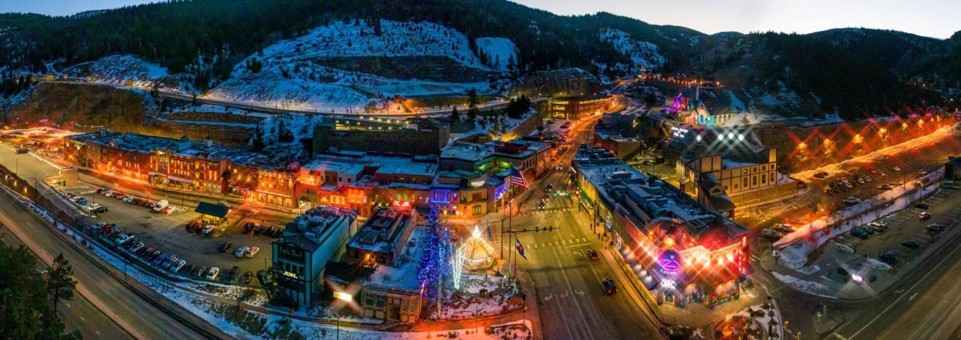 Aerial view of a brightly lit town nestled in a valley, surrounded by mountains. Lights from buildings and streets illuminate the night.