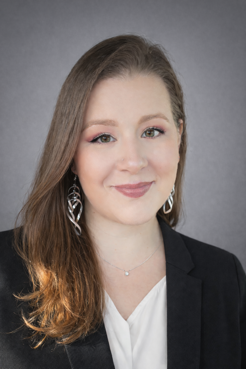 Woman with long brown hair smiles, wearing a black shirt and jacket, against a light wall.