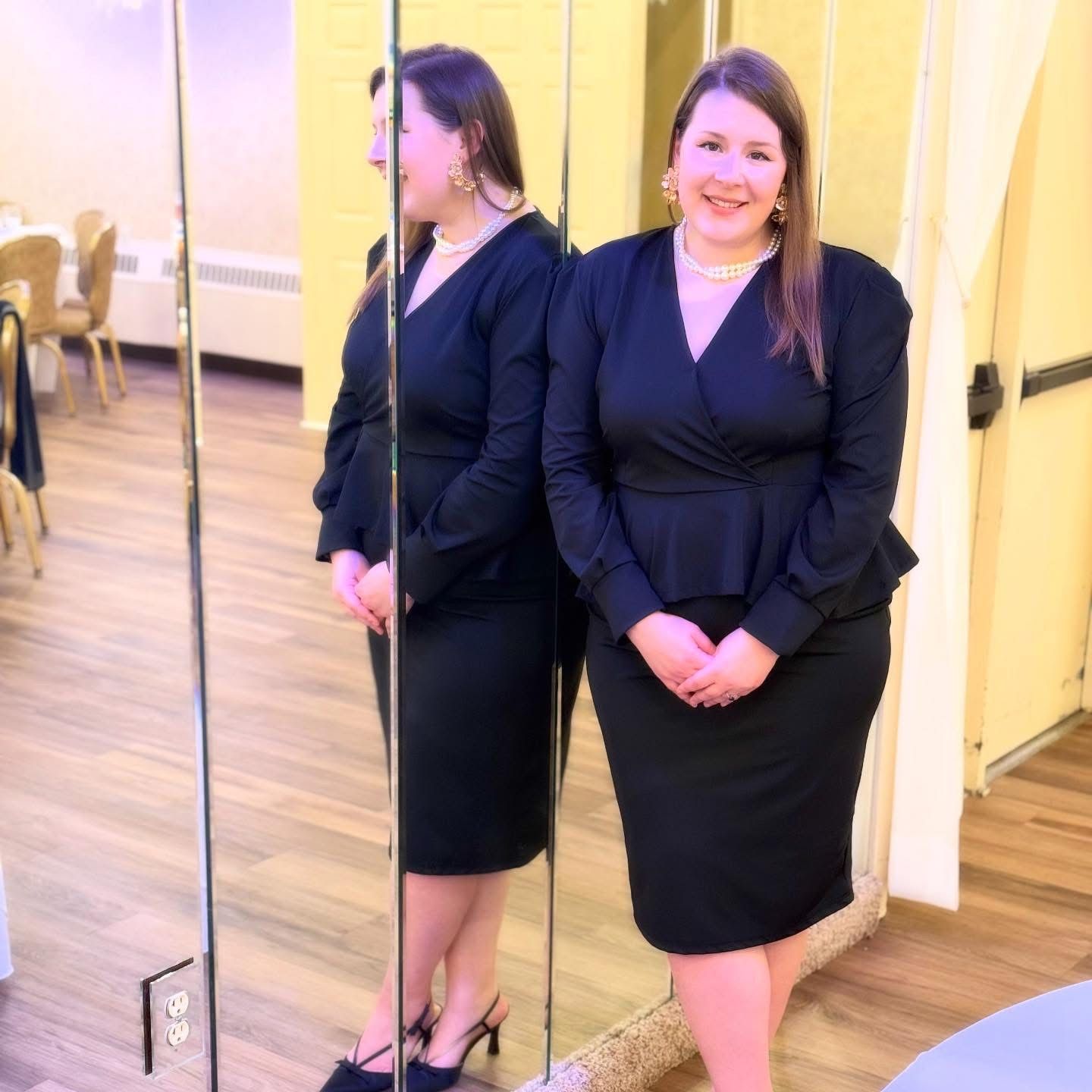 Woman in black dress and jewelry poses in front of a mirror in a room, hands clasped.