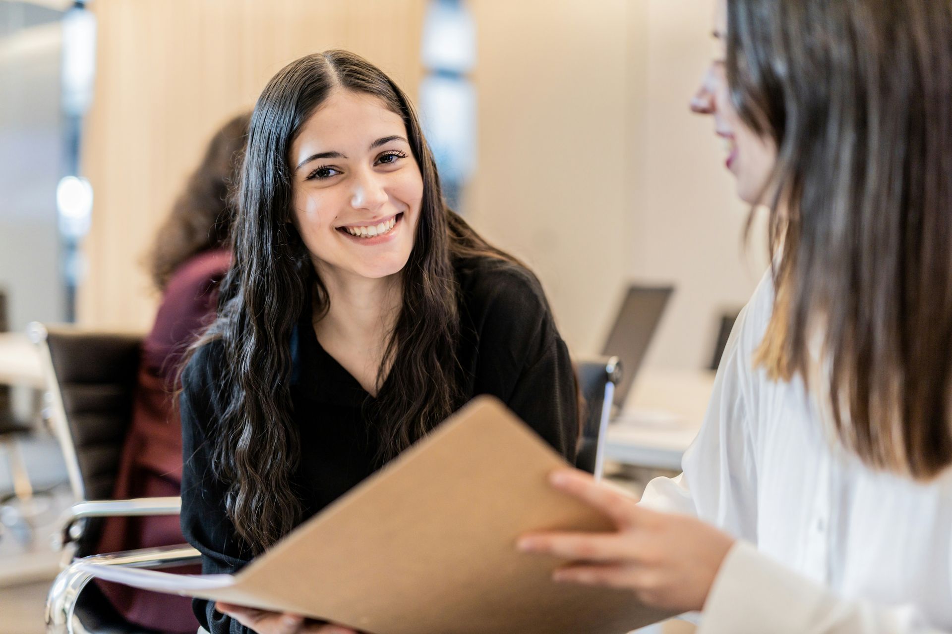 A woman is holding a folder and smiling while talking to another woman.