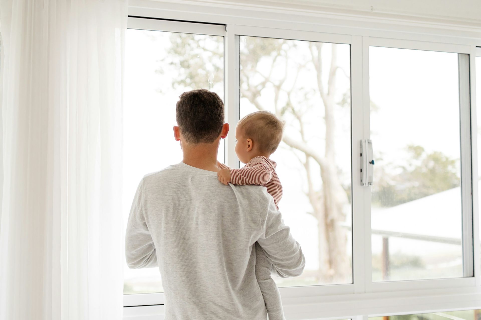 A man is holding a baby and looking out of a window.