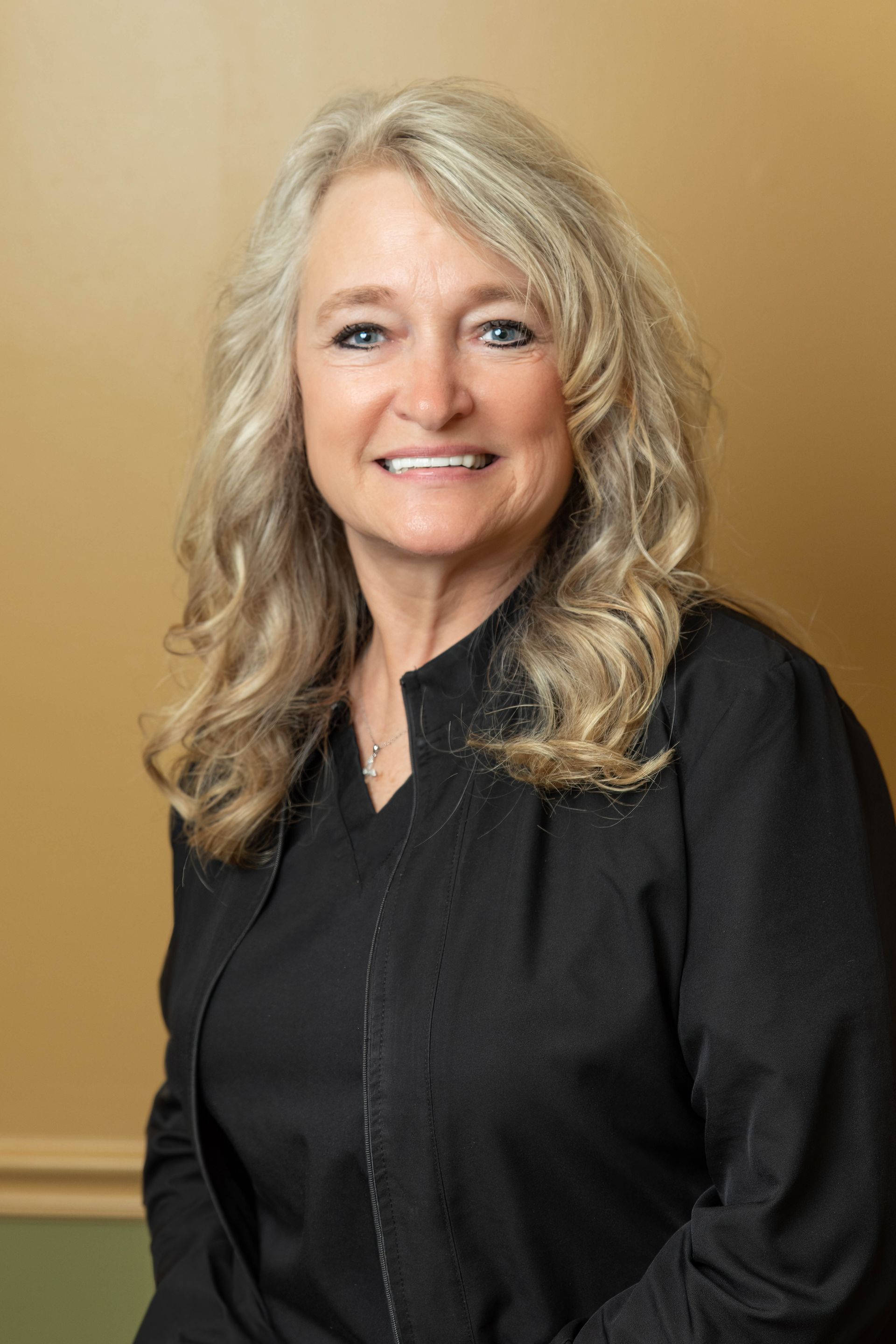 Woman with blonde wavy hair smiles, wearing a black top. Yellow wall background.
