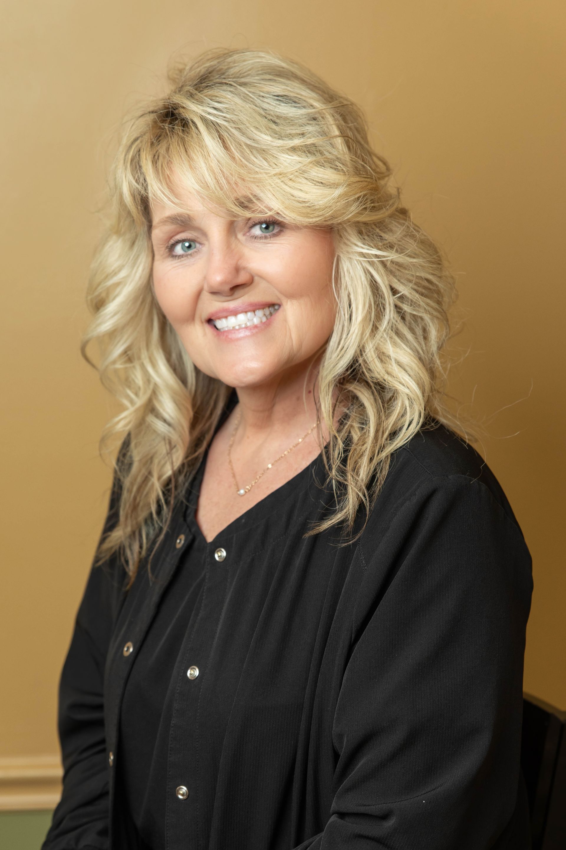 Woman with blonde hair smiling, wearing a black top, seated in front of a tan wall.