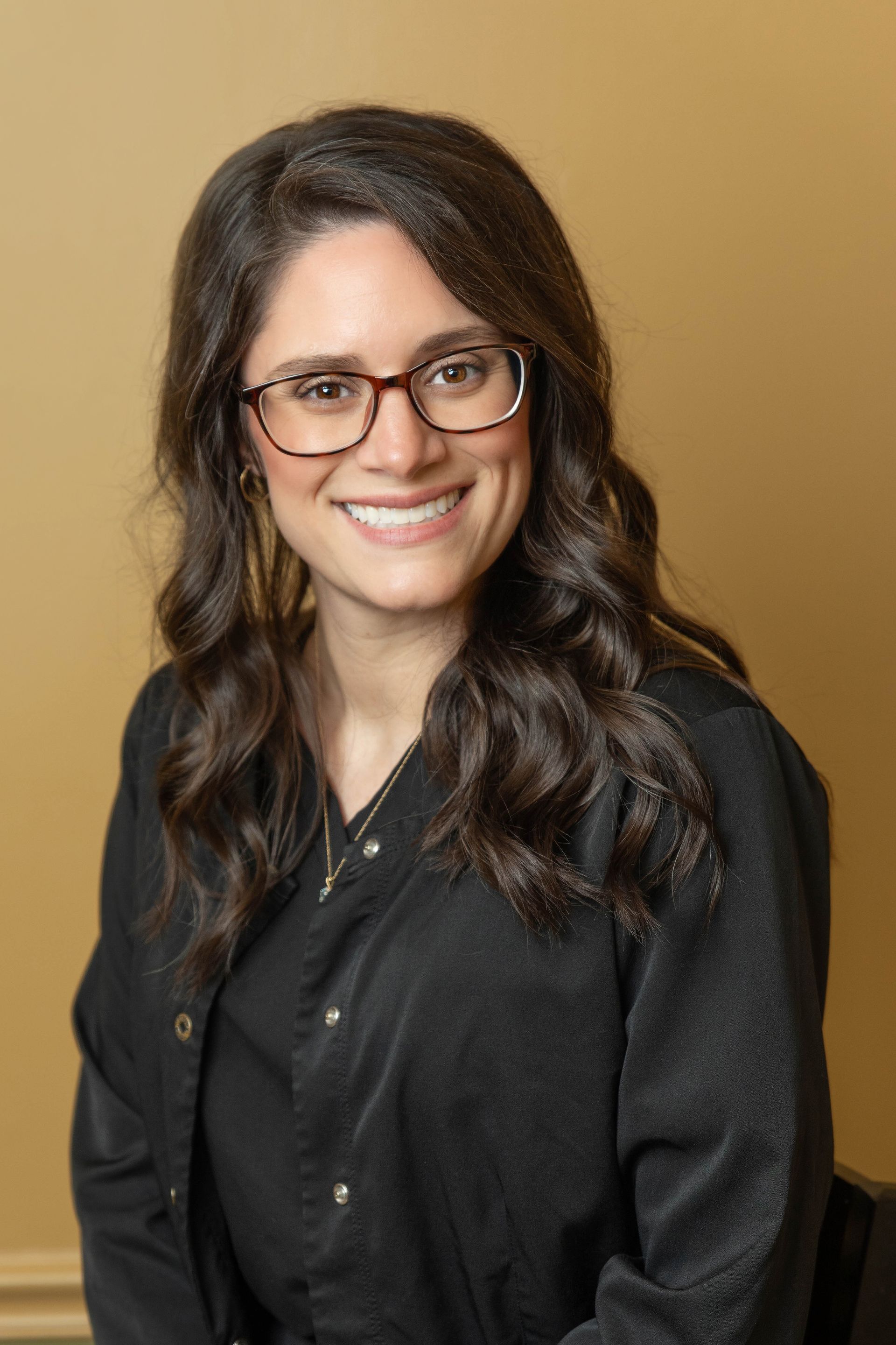 Woman wearing glasses smiles at the camera, wearing a black top against a light yellow background.