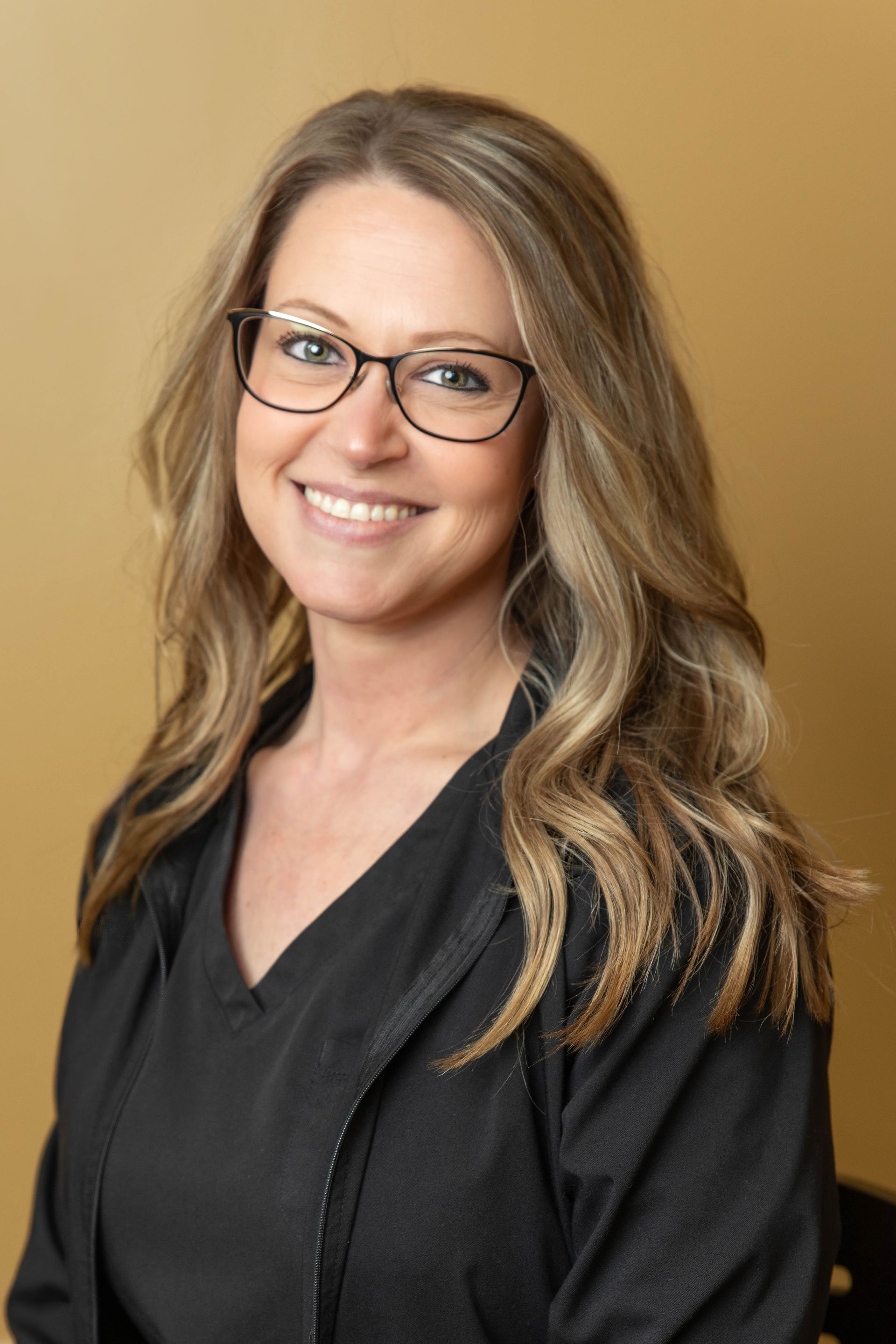 Woman wearing glasses smiles at the camera in front of a tan wall.