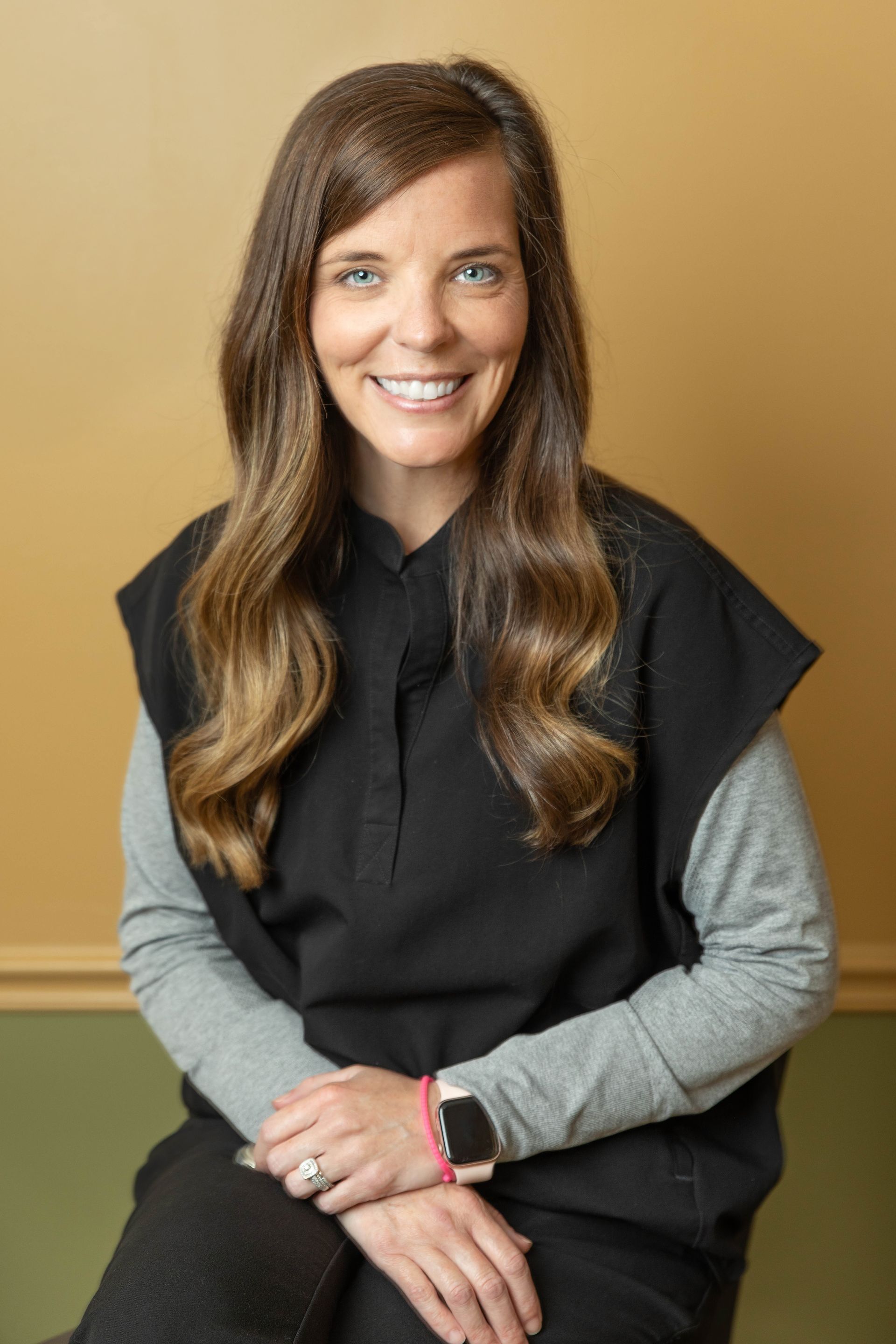 Woman in black scrubs, smiling, with a smartwatch. Sitting in a room with tan and green walls.