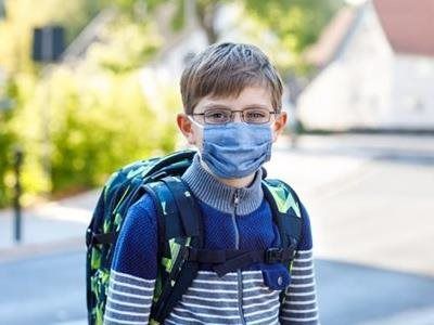 A person wearing a blue fabric face mask and glasses, carrying a green patterned backpack outdoors.
