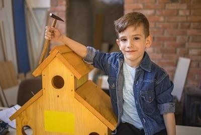 A person in a denim shirt holds a hammer over a wooden birdhouse in a workshop.