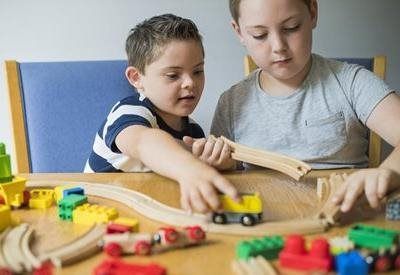 Two people sit at a wooden table building a wooden train track and playing with colorful toy train cars and blocks.