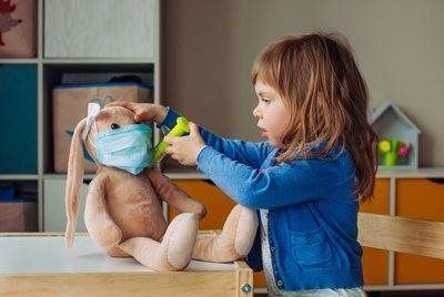 A child in a blue sweater plays doctor, holding a toy tool to a stuffed rabbit wearing a blue medical mask.