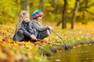 Two children in coats and hats crouch by a pond in an autumn forest, dipping sticks into the water.