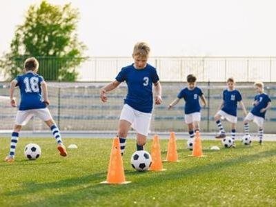 Children in blue jerseys and white shorts practice soccer drills with orange cones on a grass field.