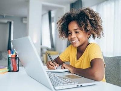 A student in a yellow shirt sits at a desk, smiling while writing in a notebook during a lesson on a laptop.