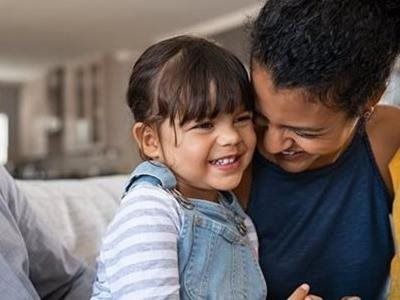 A smiling young child in a striped shirt and denim overalls sits beside a woman indoors.