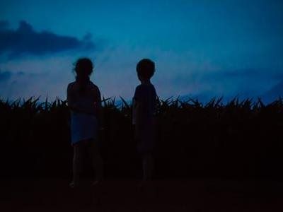 Silhouetted figures of two children standing before a field against a blue dusk sky.