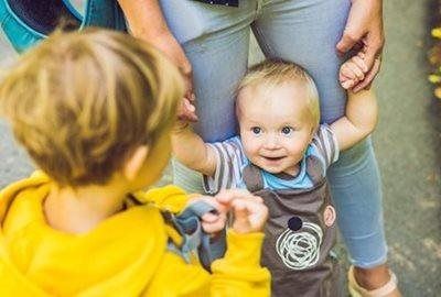 A person wearing light jeans holds the hands of a small child in gray overalls, while a child in a yellow hoodie watches.