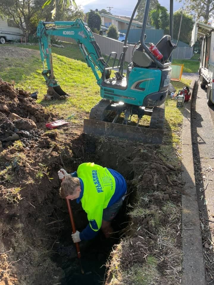 Worker in high-vis vest digging trench near a small excavator on a grassy area next to a road. — All Stars Plumbing In Dapto, NSW