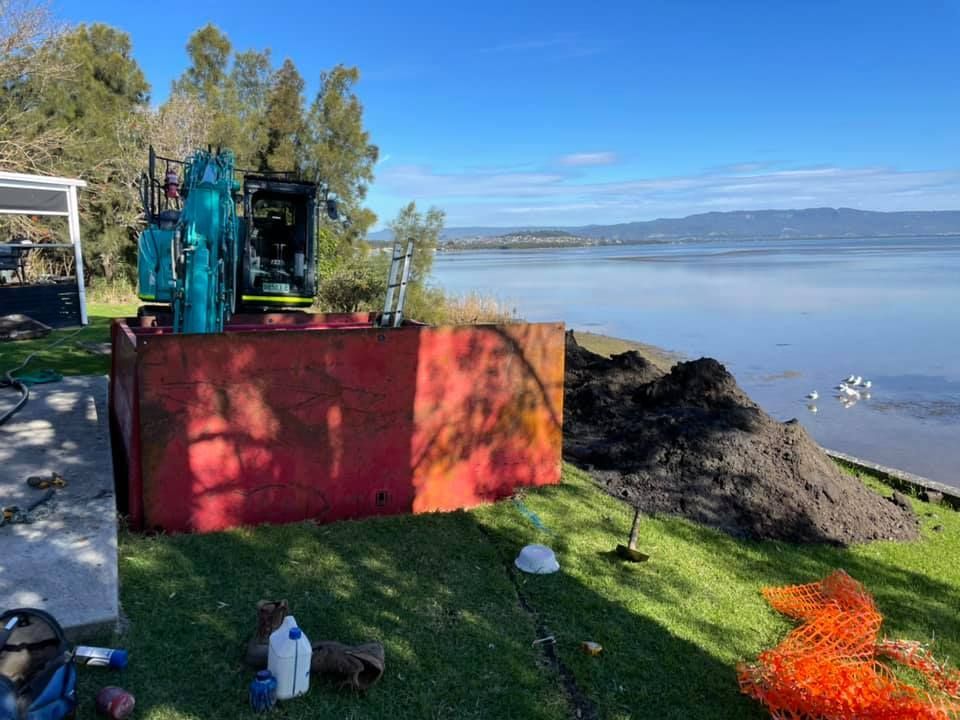 An excavator next to a red metal structure on a grassy bank by a lake under a blue sky. — All Stars Plumbing In Barrack Heights, NSW