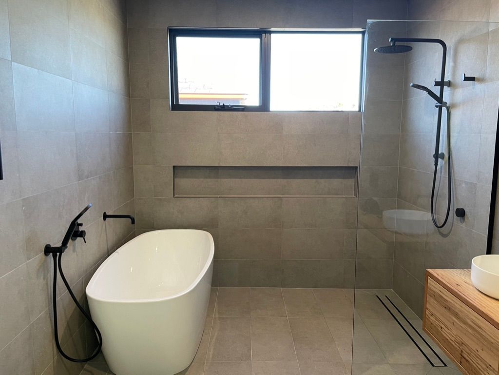 Modern bathroom with freestanding tub, shower, window, and wood vanity. Gray tile and black fixtures. — All Stars Plumbing In Dapto, NSW