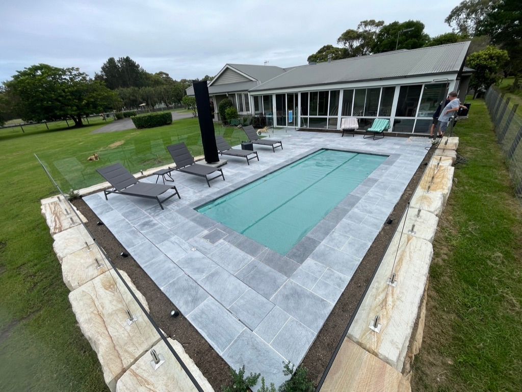 Pool with lounge chairs and house, surrounded by stone and grass. — All Stars Plumbing In Dapto, NSW