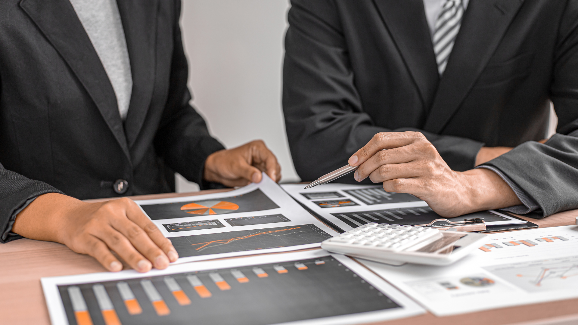 Two people in suits reviewing financial charts, with calculator and pen on desk.