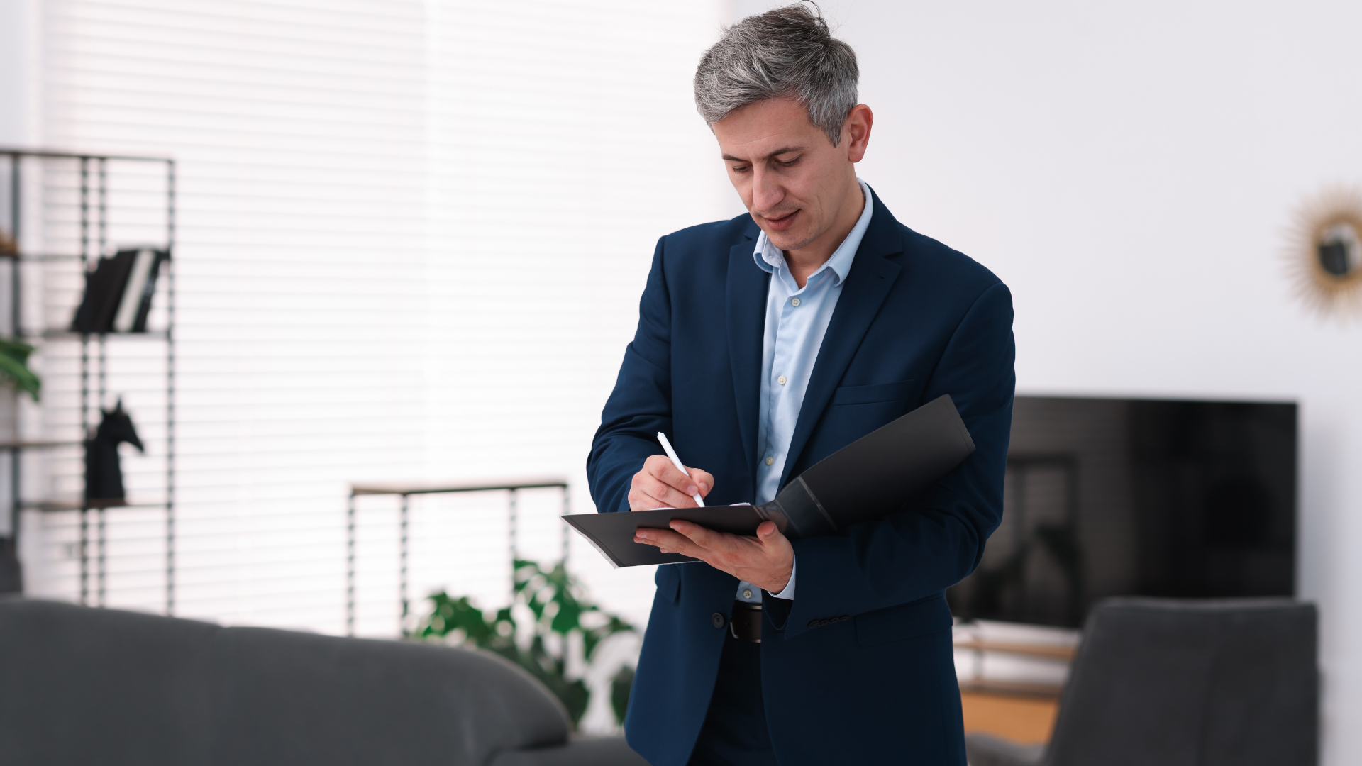Man in blue suit writing on a clipboard in a well-lit living room.