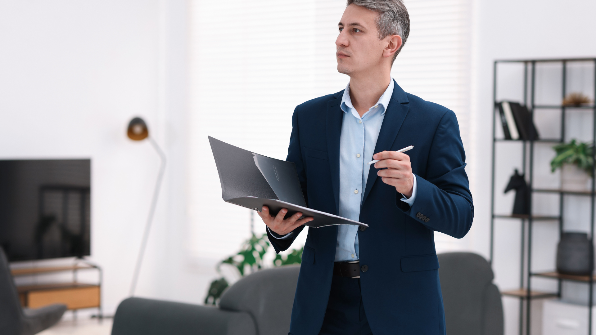 Man in a suit holding a laptop and pen, looking off to the side in a modern living room.