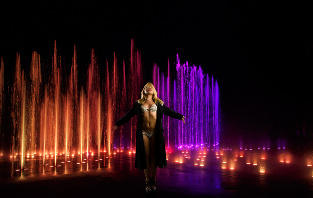 A woman in a bikini is standing in front of a fountain at night.