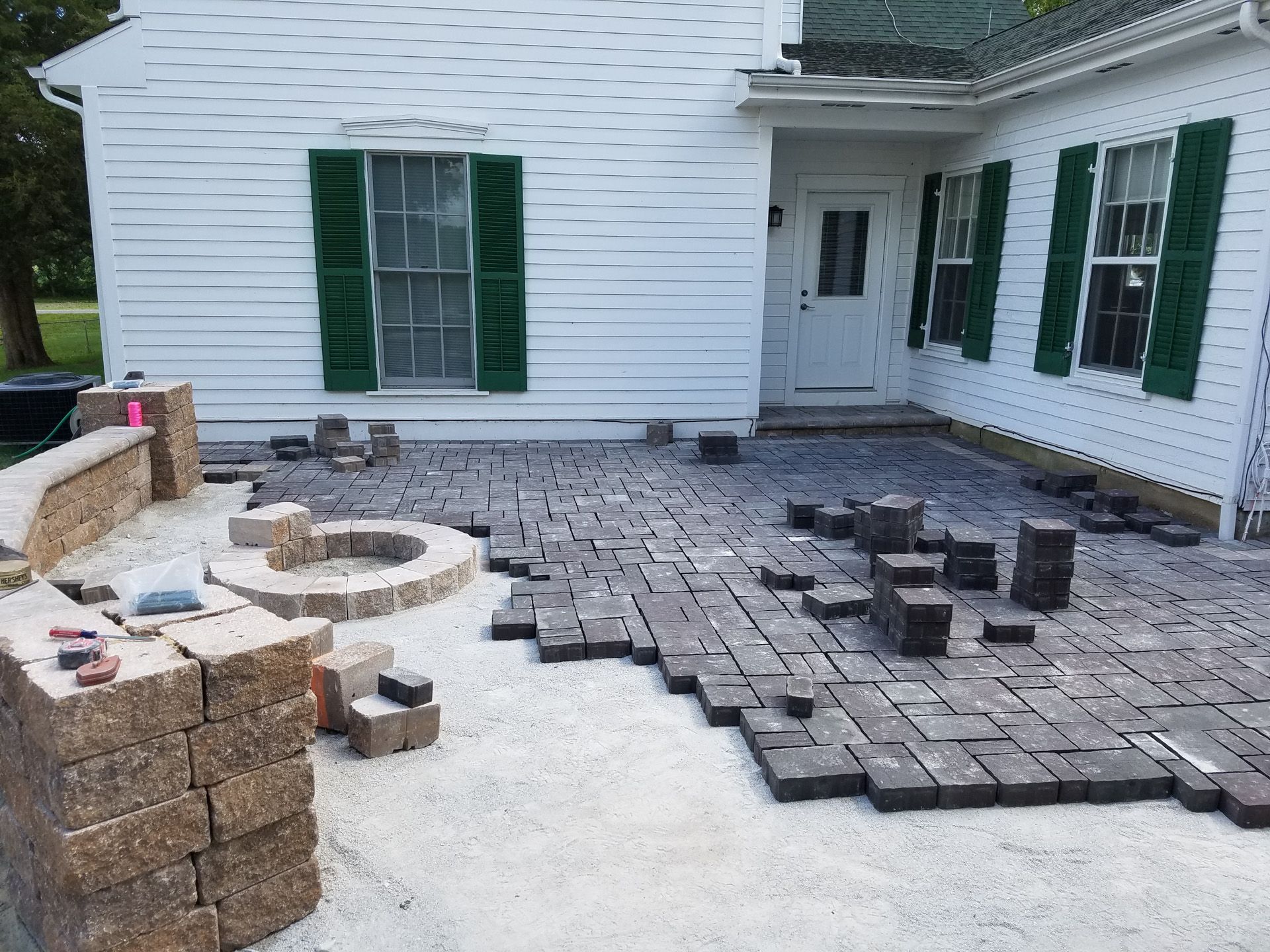 A brick patio is being built in front of a white house with green shutters.