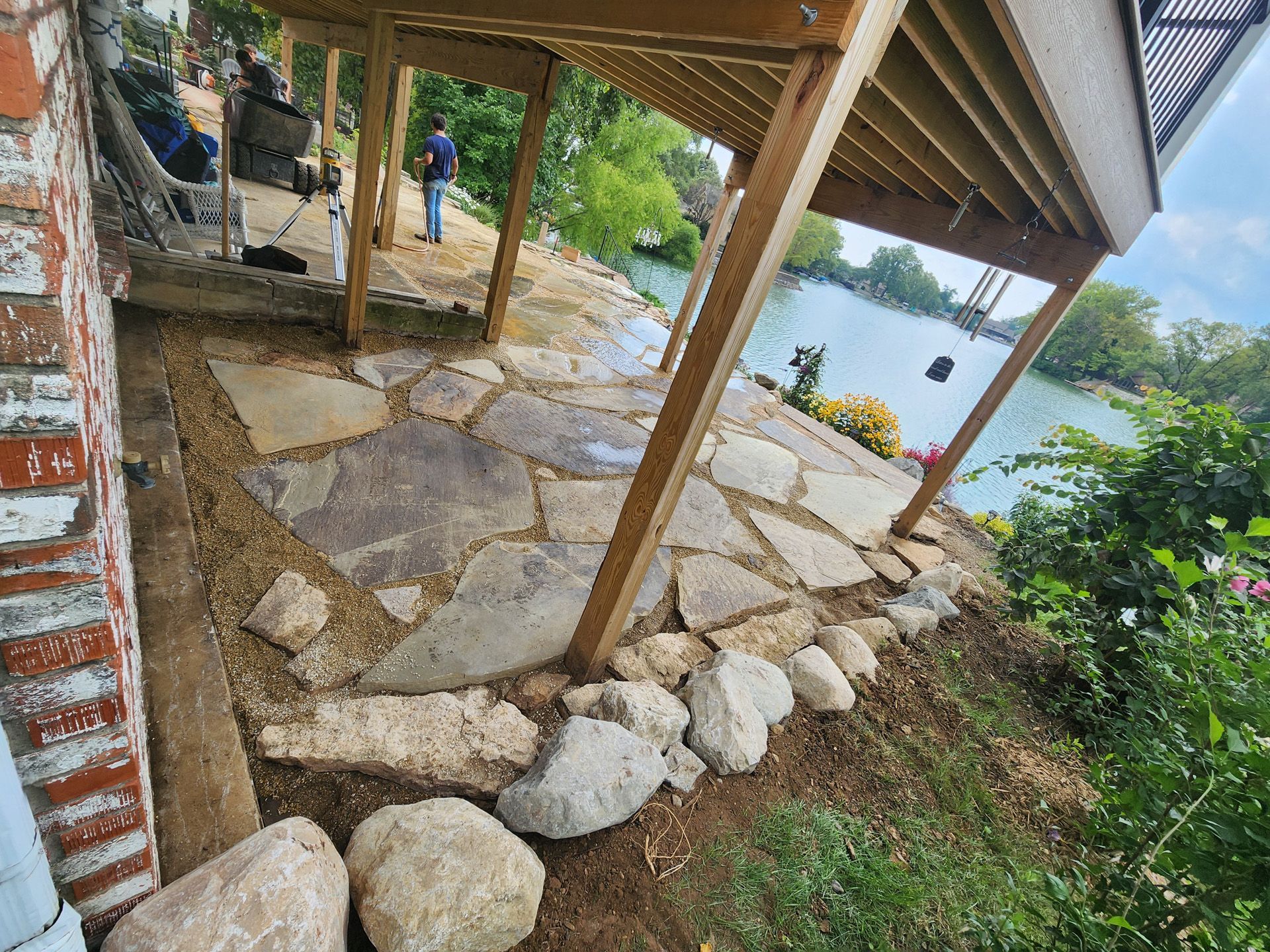 A man is standing under a wooden deck overlooking a lake.
