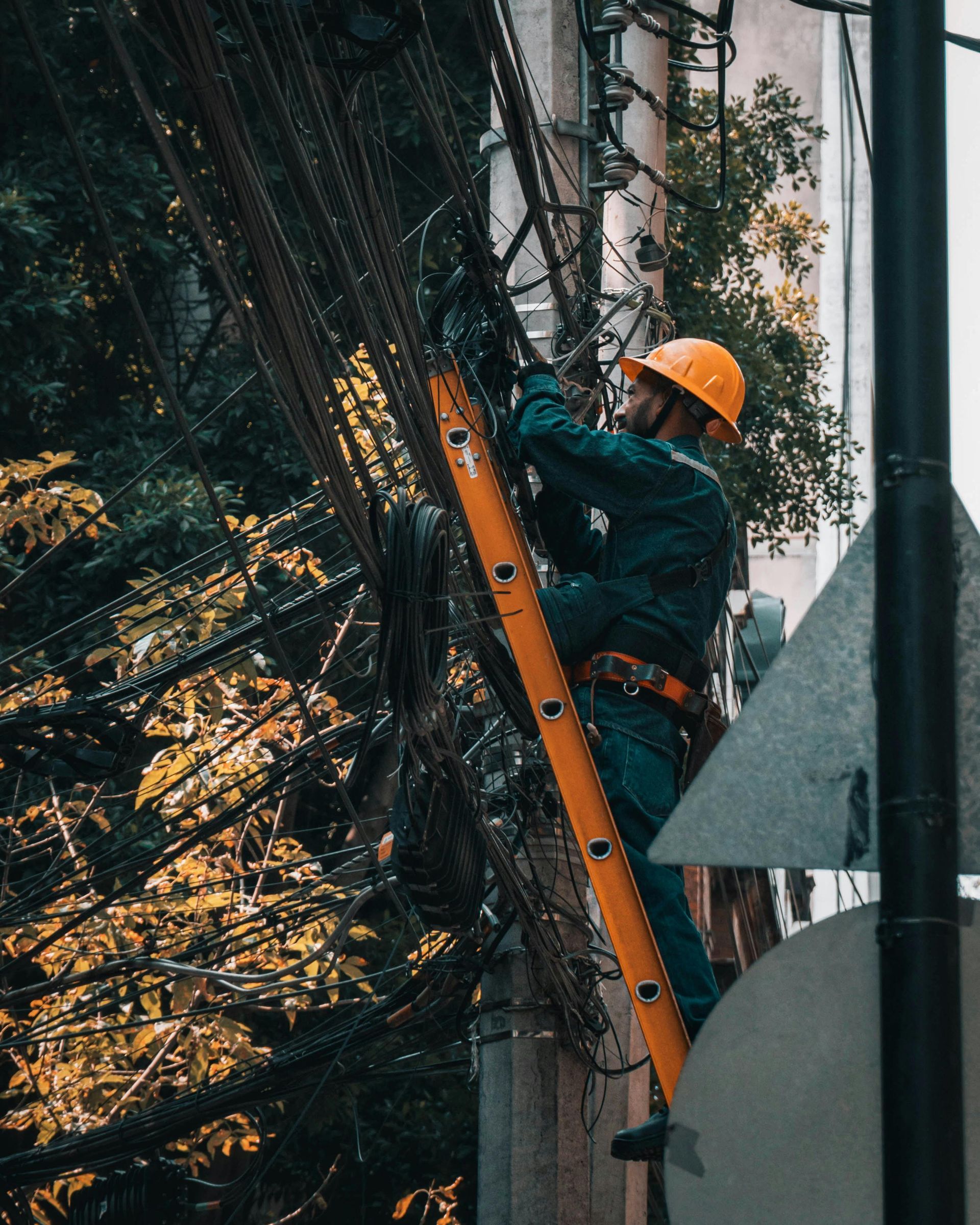 Electrician in an orange hard hat on a ladder, working on power lines attached to a utility pole.