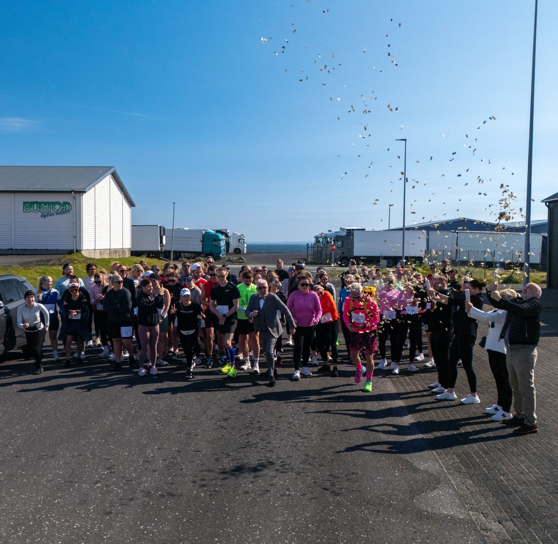 Hópur hlaupara safnaðist saman við ráslínu utandyra, með byggingar og bláan himin í bakgrunni.