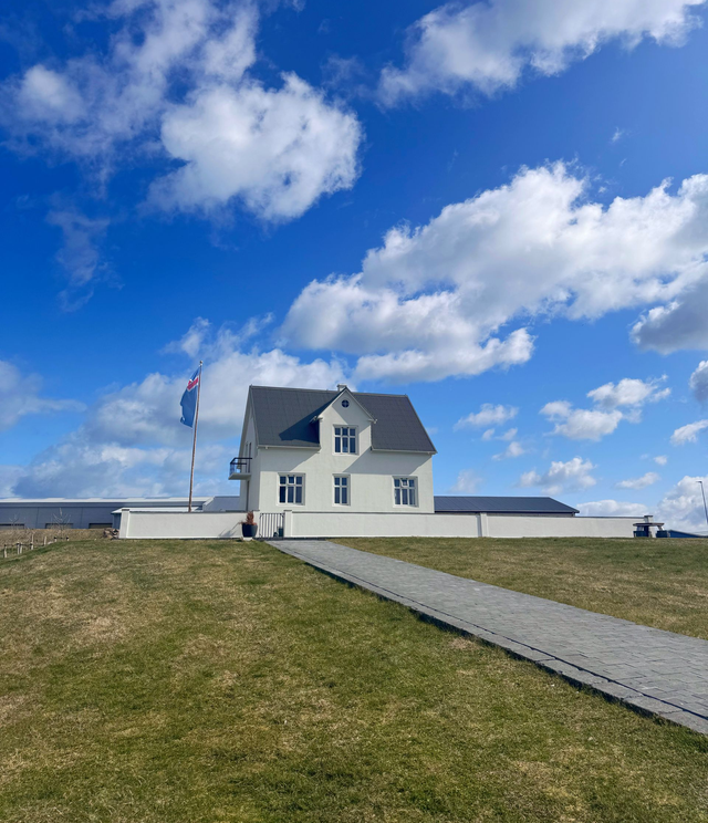 Hvítt hús á grasi vaxinni hæð með malarstíg, undir skærbláum himni með dreifðum skýjum.