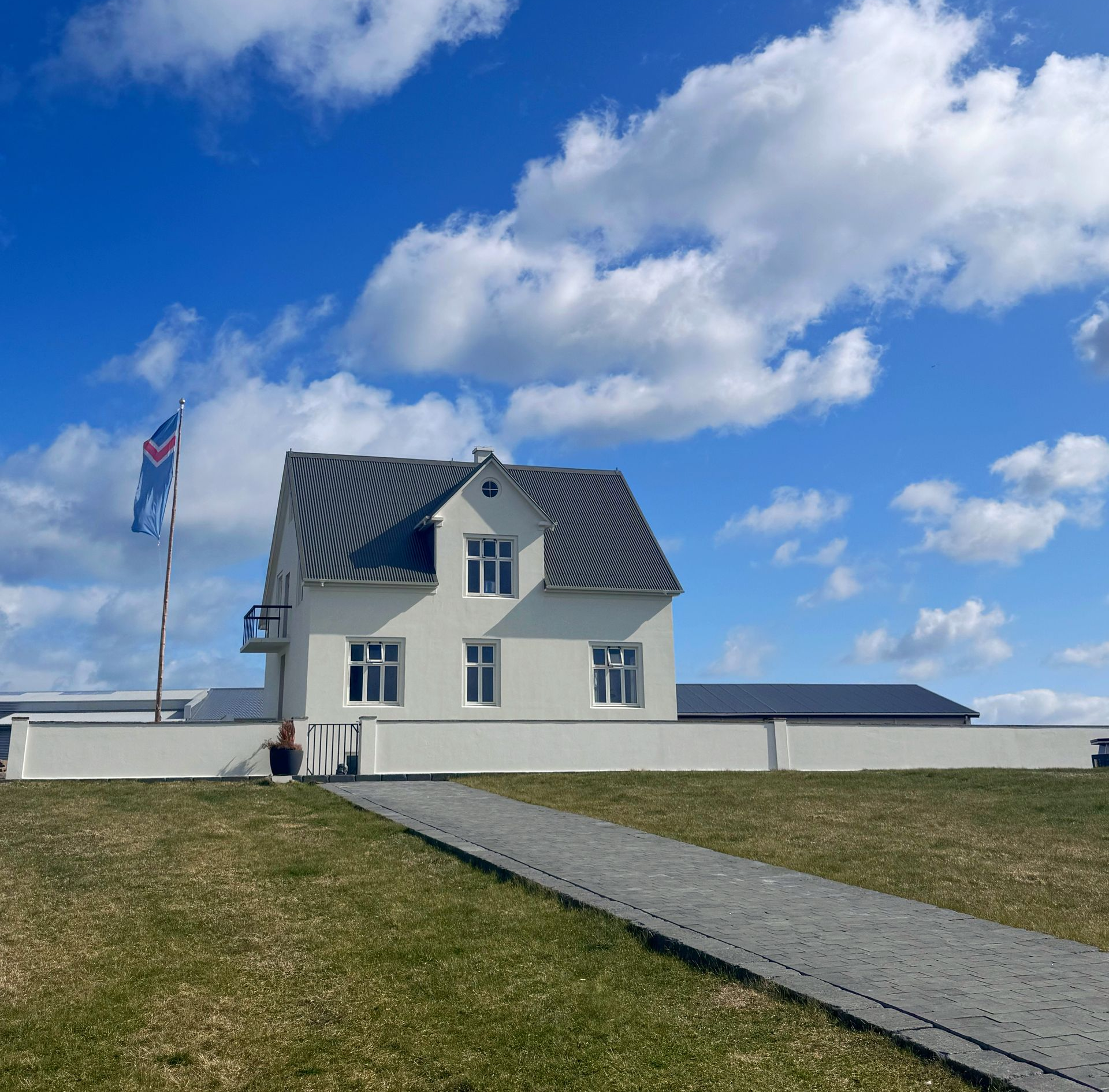 Hvítt hús með gráu þaki og fána á grasi vaxinni hæð undir bláum himni með skýjum