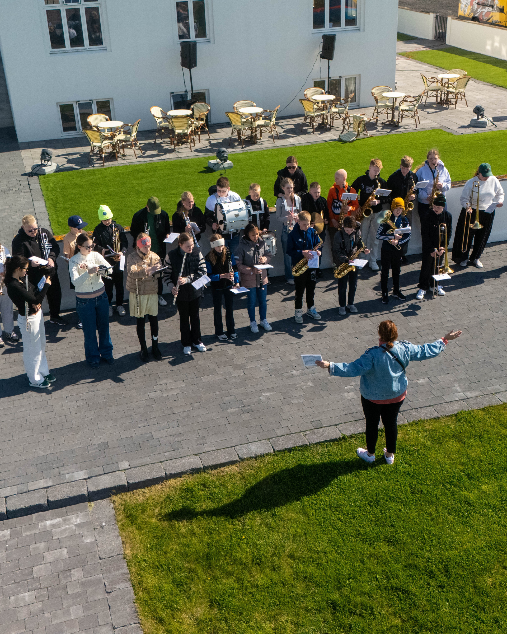 Hópur fólks stendur úti á malarstíg og snýr að manneskju með útréttar hendur á grasflöt.