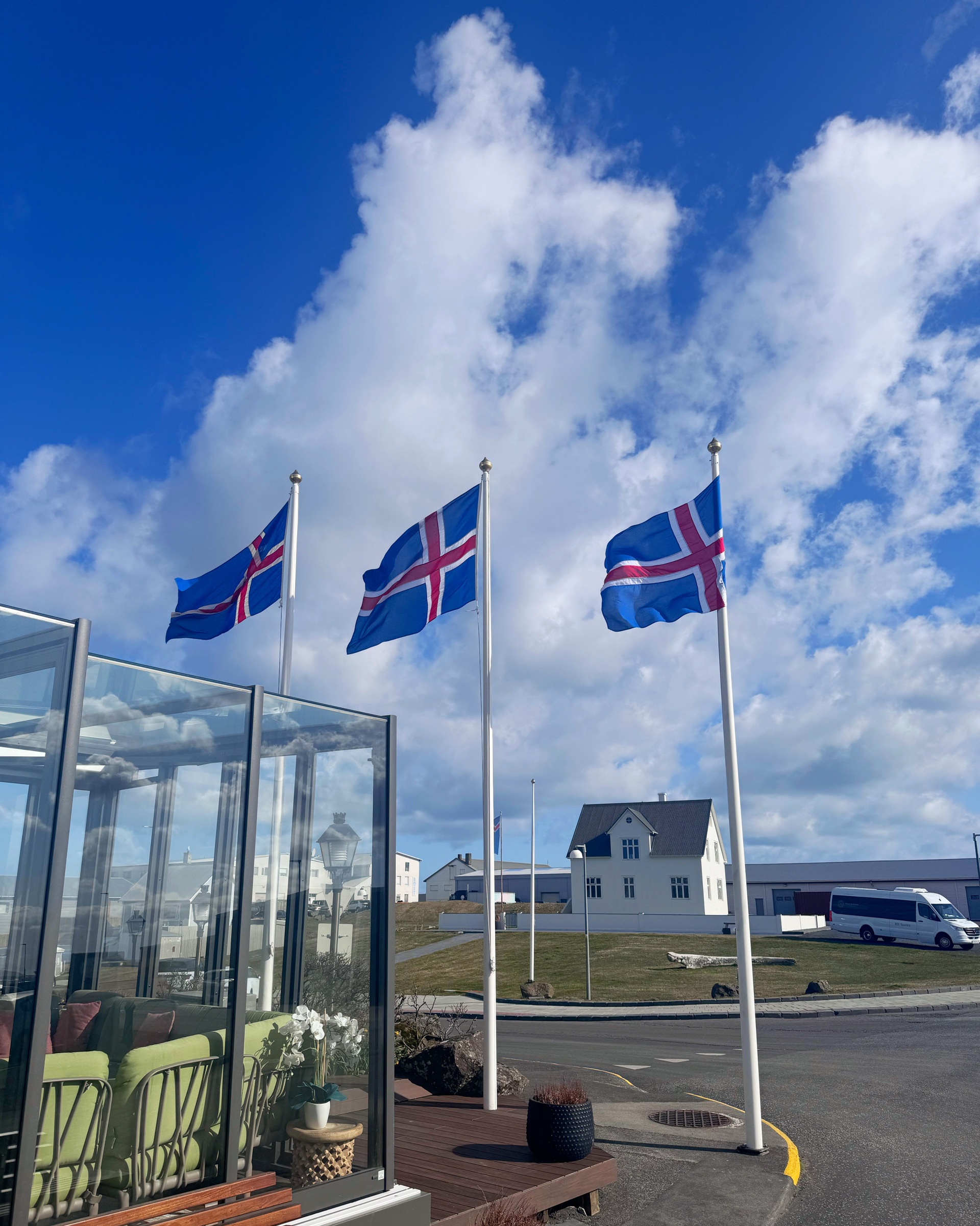 Þrír íslenskir fánar á stöngum við hliðina á glergróðurhúsi undir bláum himni með skýjum