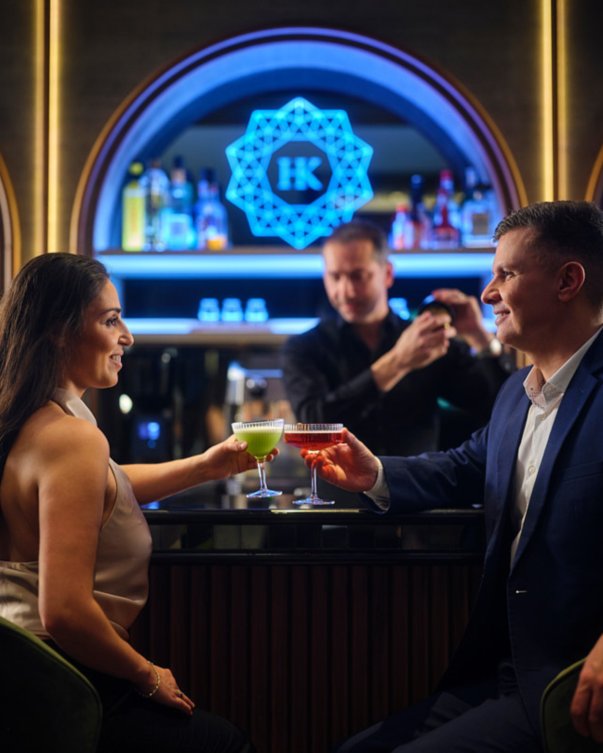 Tveir menn drekka kokteila á dauflýstum bar, með barþjón og blátt neonmerki á bak við sig.