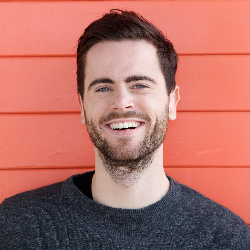A man with a beard is smiling in front of an orange wall.