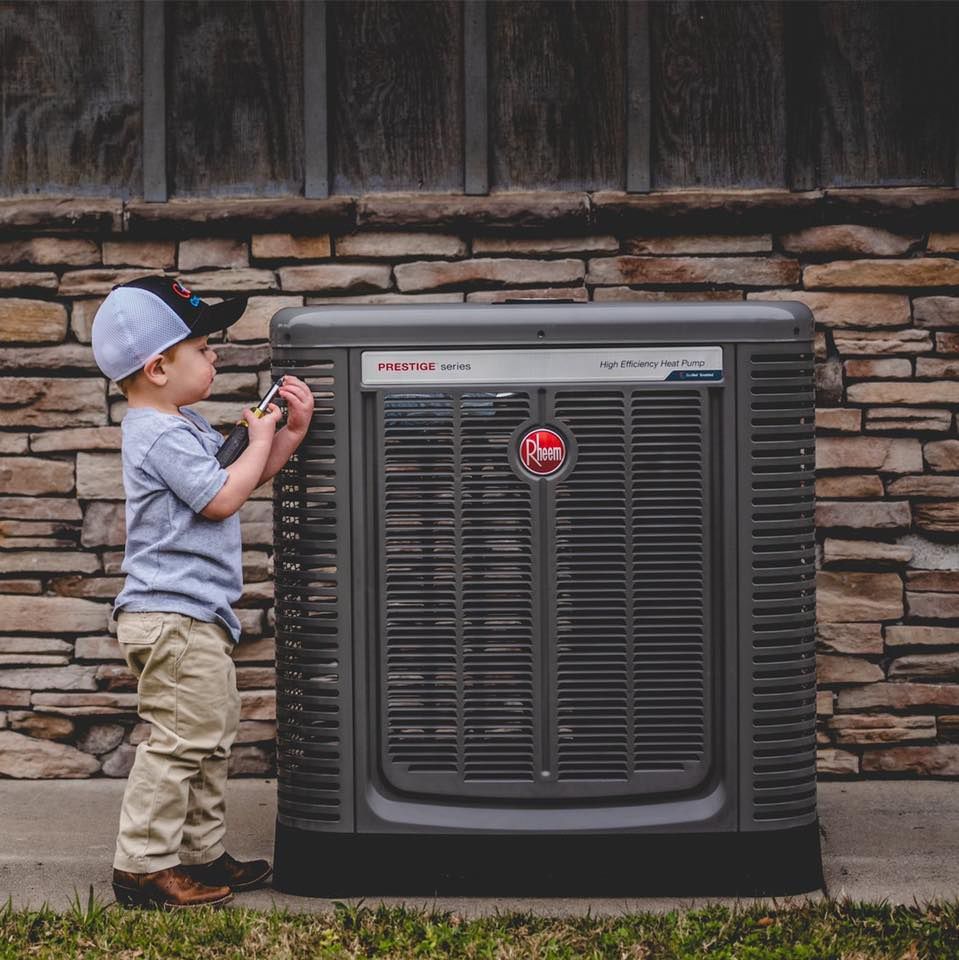 HVAC technician kneels on a rooftop, inspecting an air conditioning unit with gauges and tools under a sunny sky.