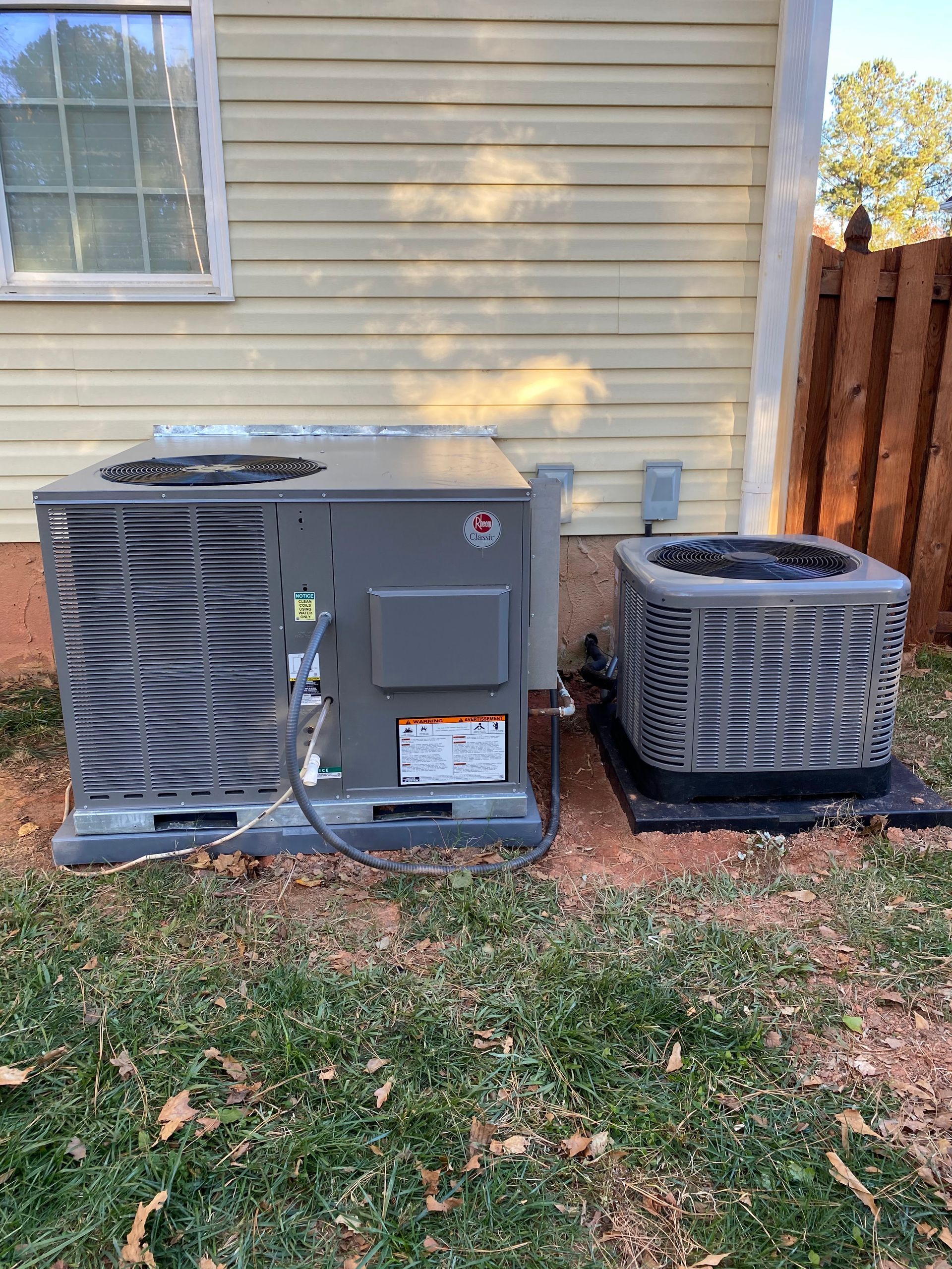 Two air conditioners are sitting next to each other on the side of a house.