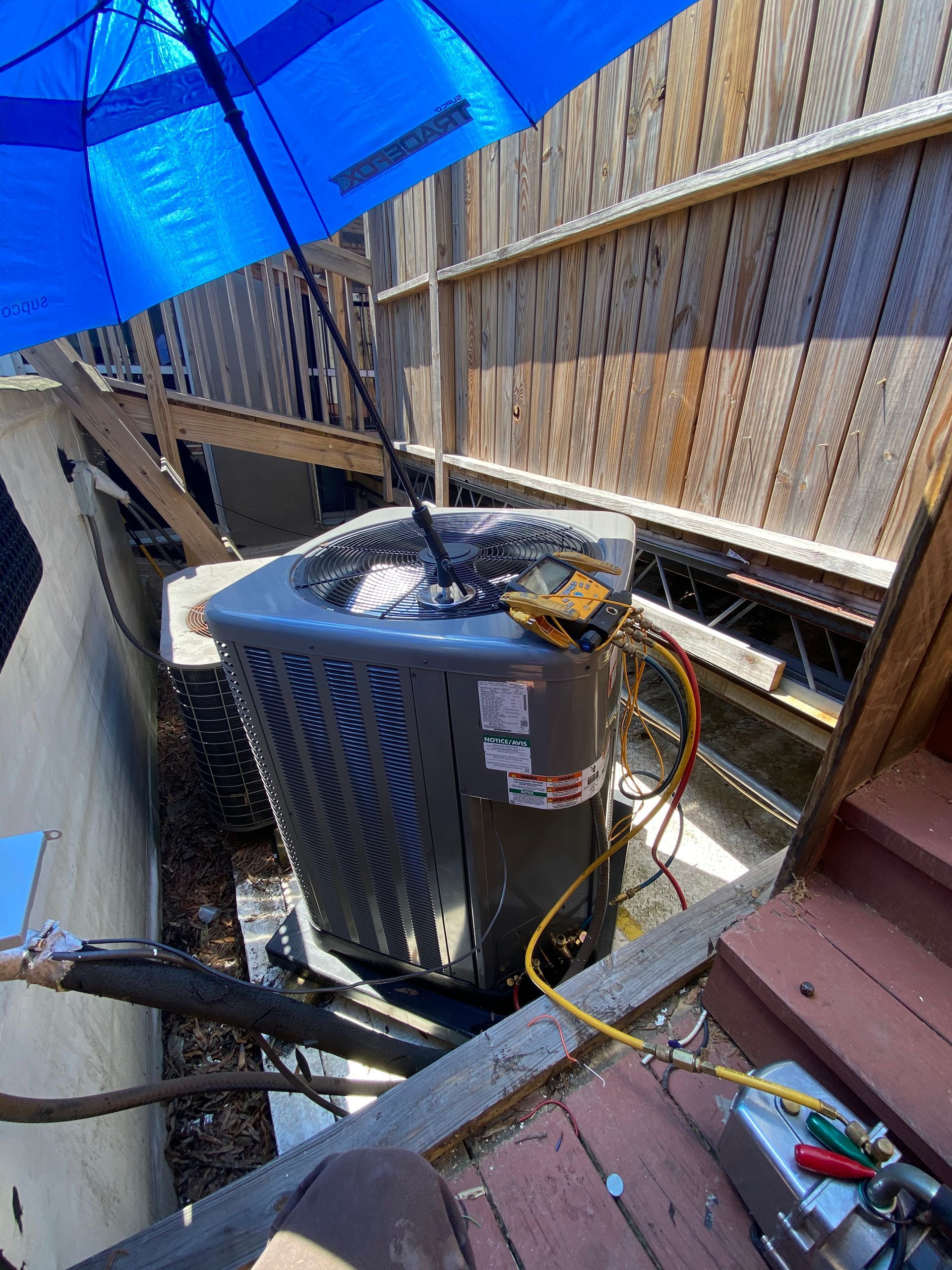 An air conditioner is being repaired on a wooden deck under an umbrella.