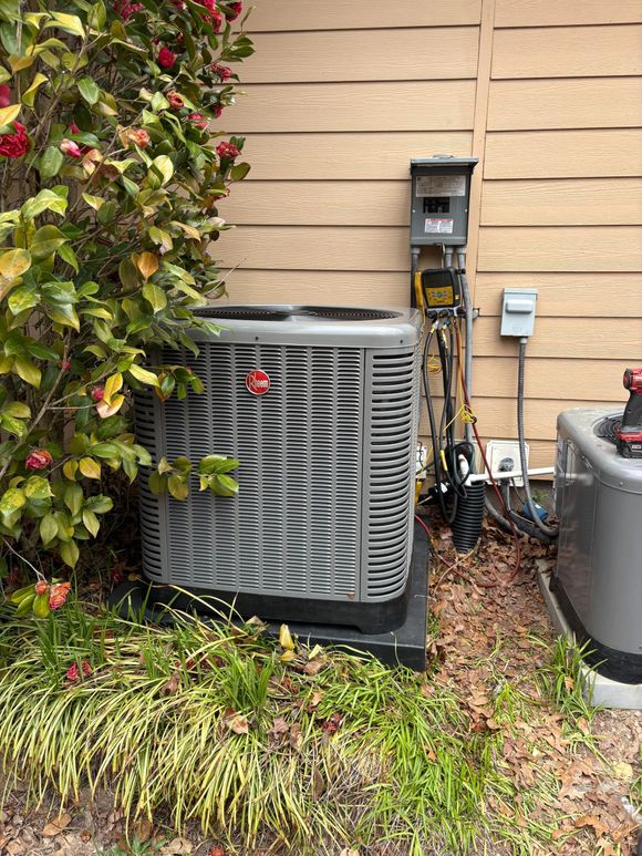 Two grey outdoor HVAC condenser units sit on the ground next to a brick building wall, surrounded by dry leaves.