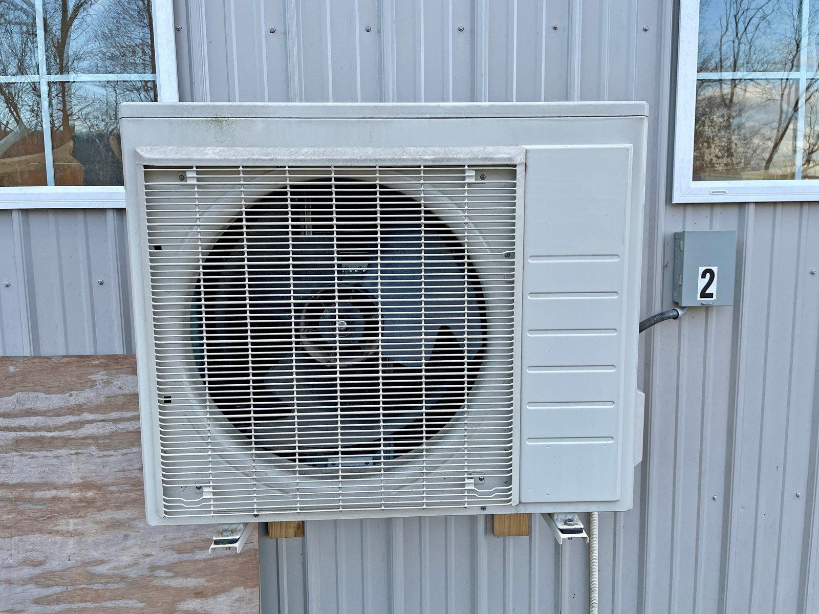 Outdoor AC unit mounted on a gray building with an electrical box.