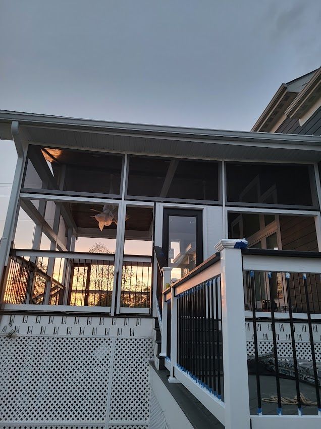 Screened porch with white railing, black accents, and a dark door. The setting is outdoors under a gray sky at dusk.