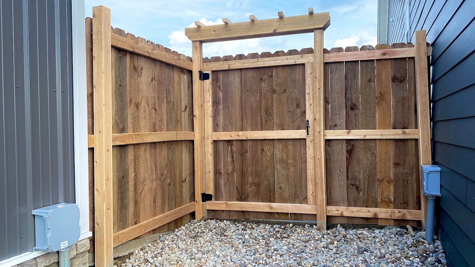 Wooden fence with gate, gravel floor, against a blue house and a metal wall, sunny day.