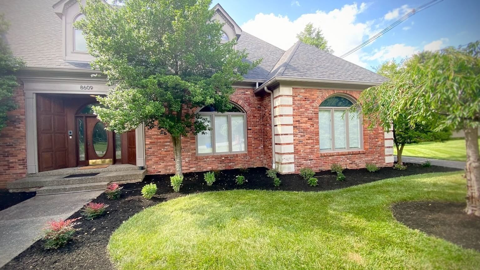 A two-story brick house with a circular turret. Green lawn, garden beds, and trees adorn the front yard.