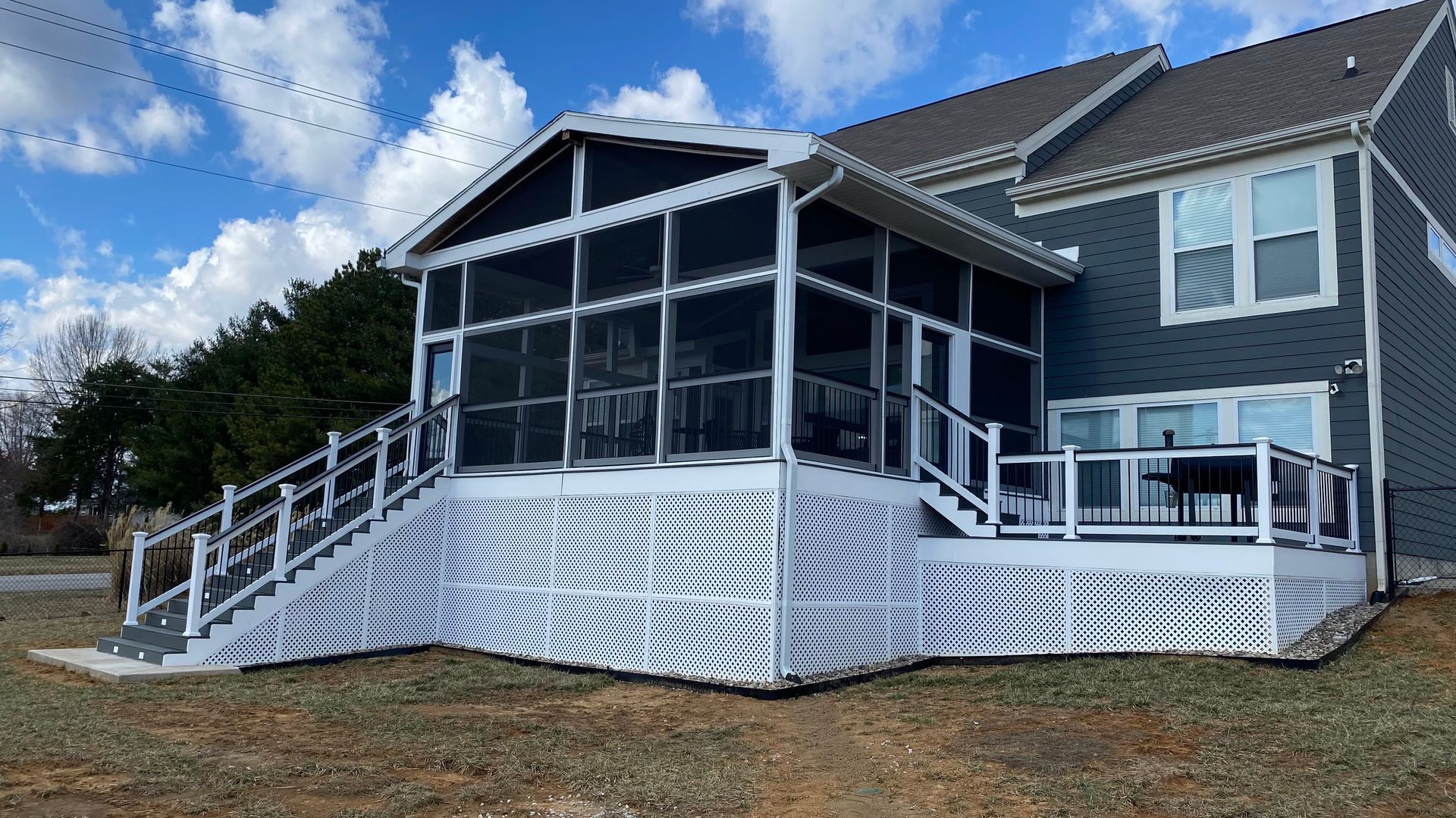 Screened porch and deck attached to a two-story blue house, with white railing and a side staircase, under a cloudy sky.