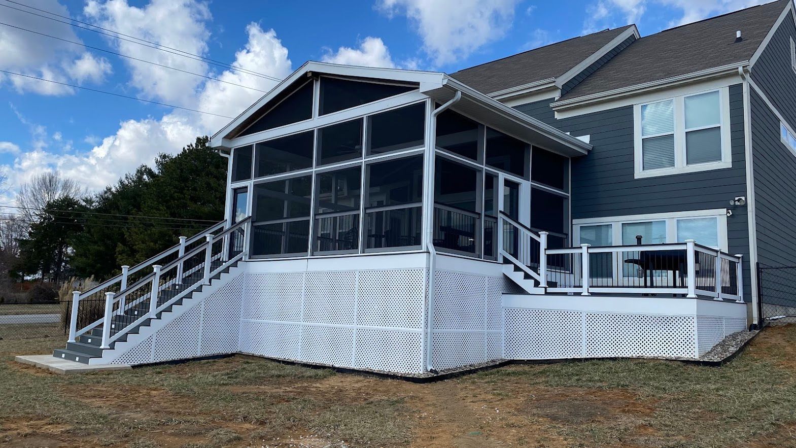White screened-in porch attached to a gray house with a white deck and railings. Blue sky with clouds.