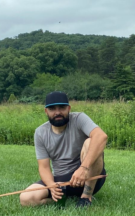 Man sitting cross-legged in a grassy field with hills and trees in the background.